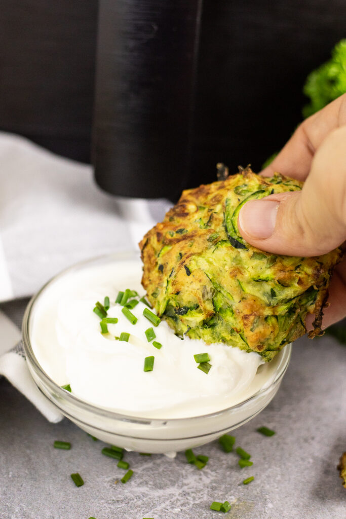 Air fryer zucchini fritter in front of an air fryer being dipped into a small bowl of sour cream topped with chives.