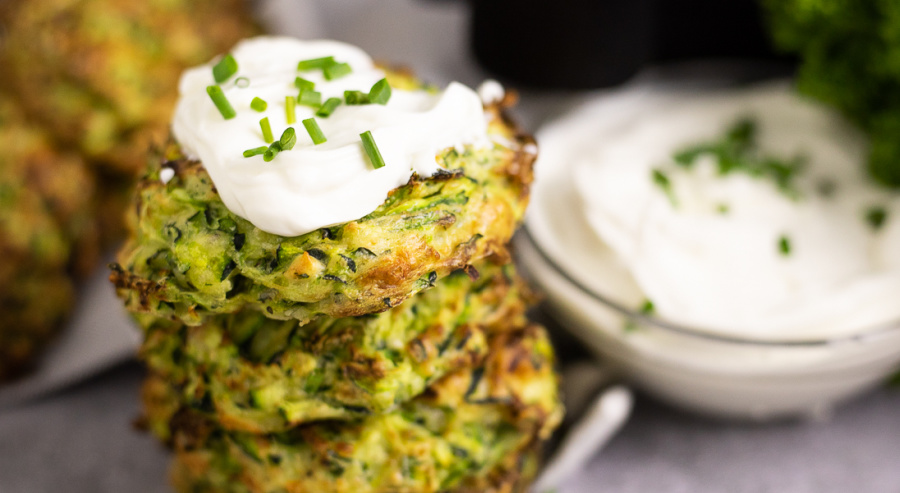 Stack of 4 zucchini fritters on a concrete backdrop with chives scattered all around. The top fritter is topped with sour cream and chopped chives.