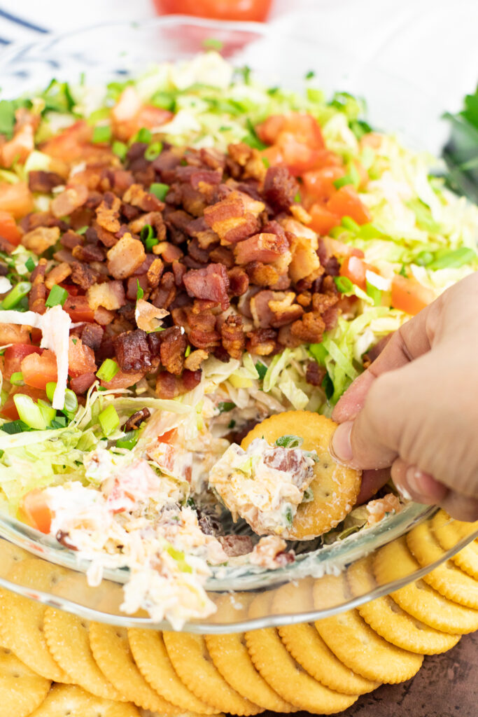 BLT Dip topped with shredded lettuce, crispy bacon, and ripe diced tomatoes with a cracker dipping into it.