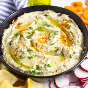 Black matte bowl filled with baba ganoush, topped with olive oil fresh parsley, and smoked paprika, surrounded by pita chips, radish slices, and carrot slices with a glass bottle of olive oil in the background