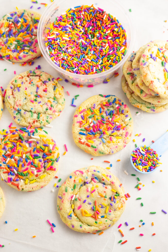 Funfetti cookies scattered over a white background with rainbow sprinkles in a cup and scattered all around