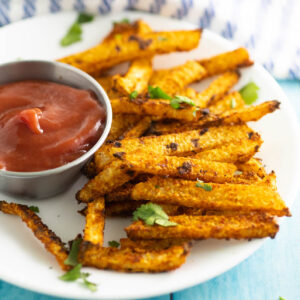 Jicama fries topped with fresh cilantro piled on a small white plate next to a small metal bowl of ketchup on a bright blue wood backdrop