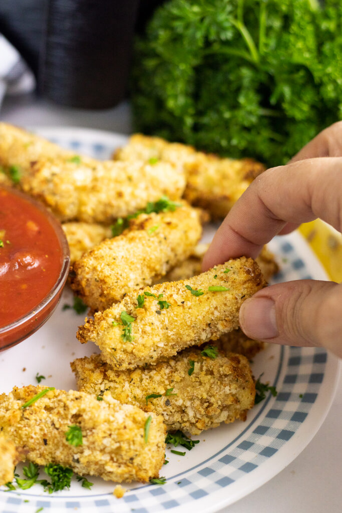 A hand picking up an air fryer mozza stick from a pile of mozzarella sticks on a plate with a bunch of fresh parsley in the background.
