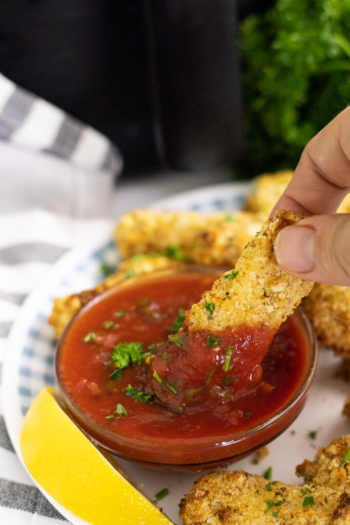 Air fryer mozzarella stick being dipped into a bowl of marinara sauce sprinkled with fresh chopped parsley next to a lemon wedge