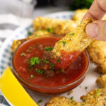 Air fryer mozzarella stick being dipped into a bowl of marinara sauce sprinkled with fresh chopped parsley next to a lemon wedge