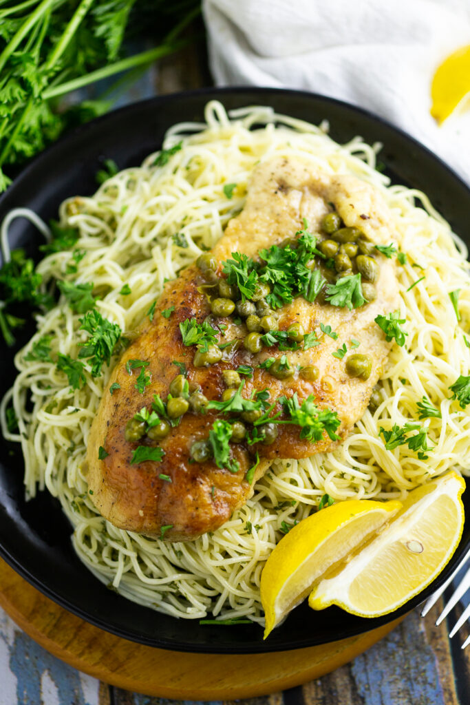 Chicken piccata on a bed of angel hair pasta topped with fresh chopped parsley next to 2 lemon wedges, a fork, and a clean linen napkin