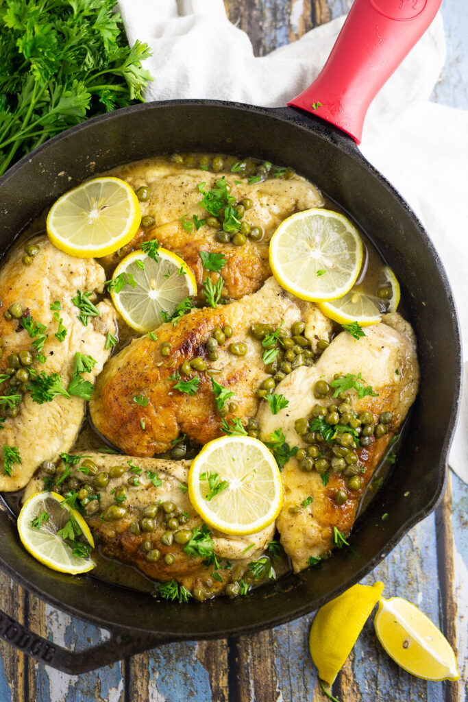 Overhead view of 5 pieces of chicken piccata in a cast iron skillet topped with lemon slices, capers, and fresh chopped parsley. Skillet is on a rustic wood background with lemon wedges and fresh parsley