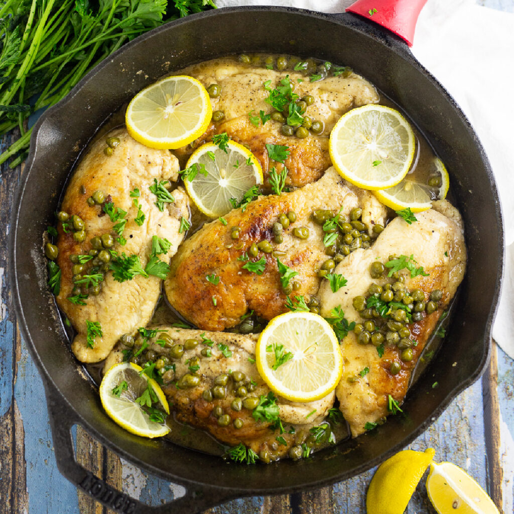 Overhead view of 5 pieces of chicken piccata in a cast iron skillet topped with lemon slices, capers, and fresh chopped parsley. Skillet is on a rustic wood background with lemon wedges and fresh parsley