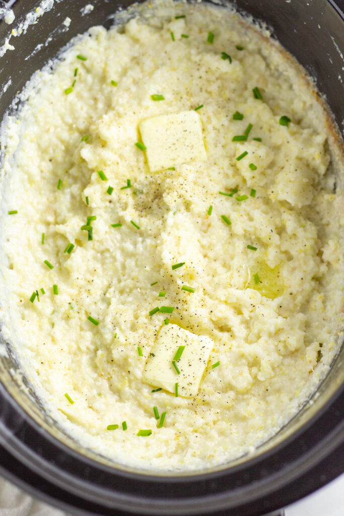 Slow cooker full of creamy grits topped with melting butter, fresh chives, and black pepper