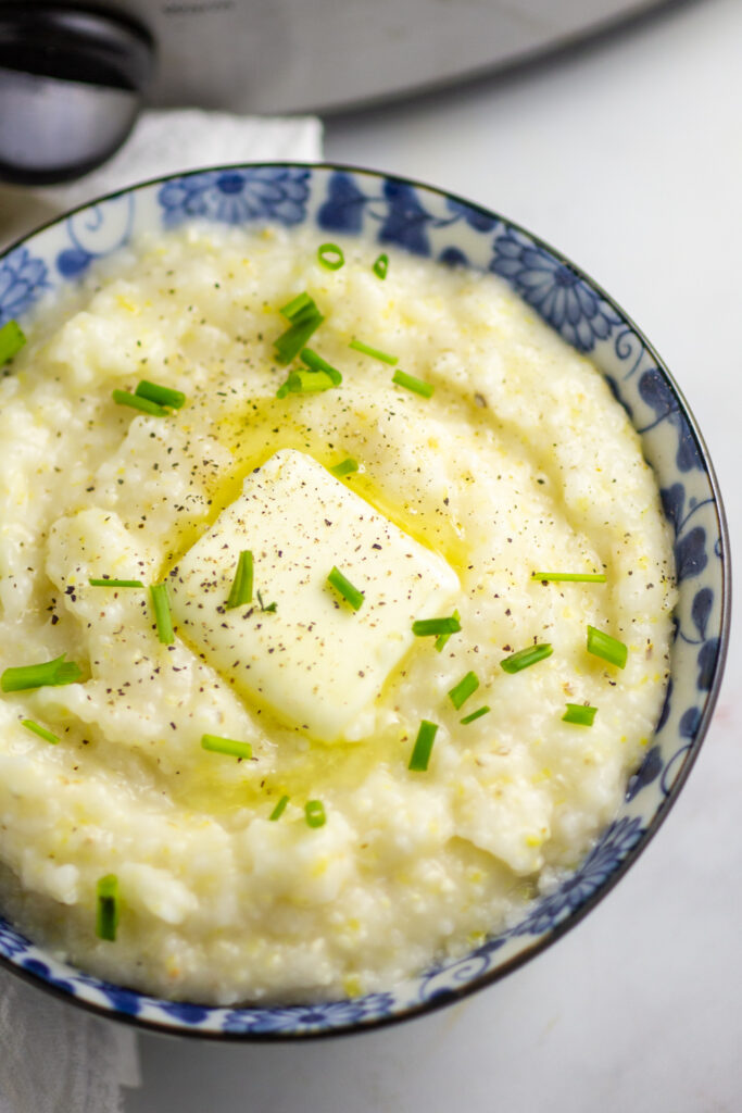 A close up of a small bowl full of crockpot grits topped with melting butter, fresh chopped chives, and black pepper,
