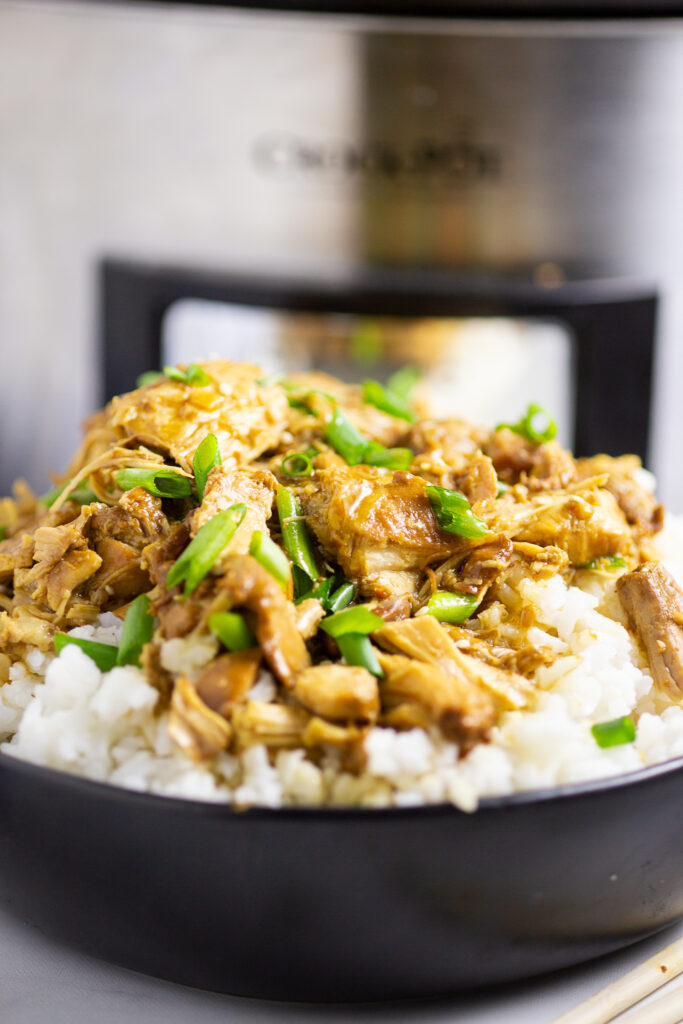 Close up side view of crockpot bourbon chicken over rice topped with sliced green onions and a stainless steel Crock Pot in the background