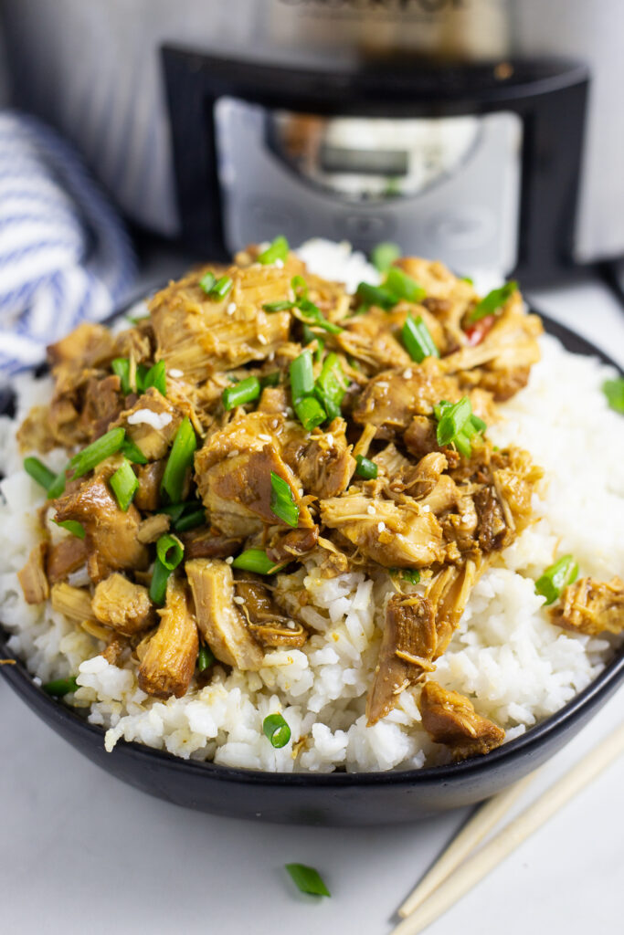 Crockpot bourbon chicken topped with sliced green onions over rice in a black matte bowl