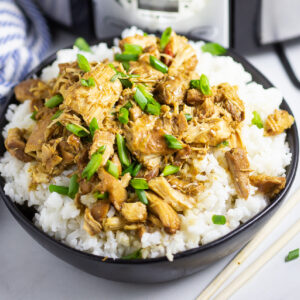 Crockpot bourbon chicken topped with sliced green onions over rice in a black matte bowl