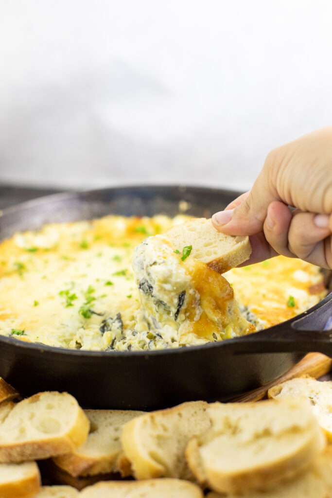 Baguette slice pulling gooey cheese and hot spinach artichoke dip from a cast iron skillet