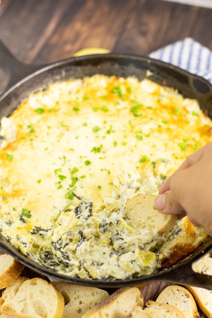 Cast iron skillet full of hot spinach artichoke dip topped with fresh chopped parsley with a baguette slice being dipped into it. 