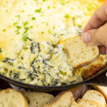 Slice of baguette being dipped into the edge of hot spinach artichoke dip in a cast iron skillet
