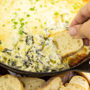 Slice of baguette being dipped into the edge of hot spinach artichoke dip in a cast iron skillet