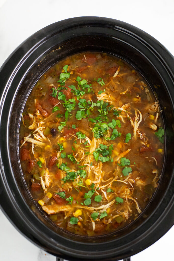 Slow cooker filled with chicken tortilla soup topped with fresh cilantro on a white marble background