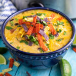 A dark blue bowl in front of a slow cooker. The bowl is full of slow cooker chicken tortilla soup with sour cream stirred in and topped with shredded cheese, tortilla strips, and fresh cilantro.