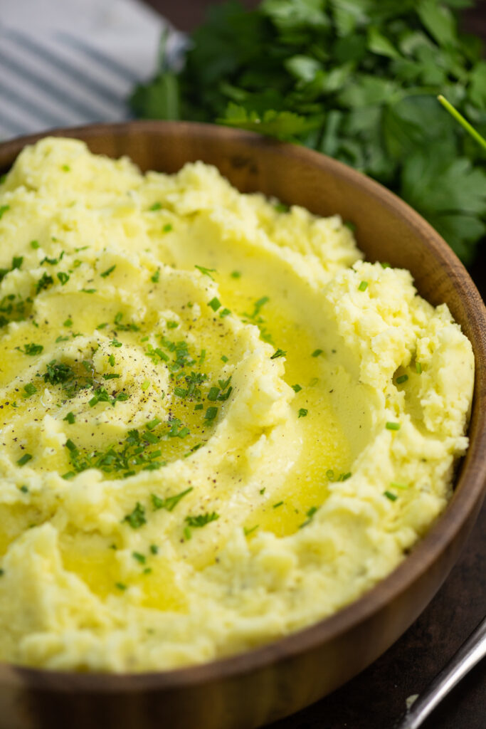 Wooden bowl full of Boursin mashed potatoes topped with melted butter and freshly chopped chives with a bunch of fresh parsley behind