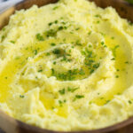 Wooden bowl full of Boursin mashed potatoes topped with melted butter and freshly chopped chives with a bunch of fresh parsley behind