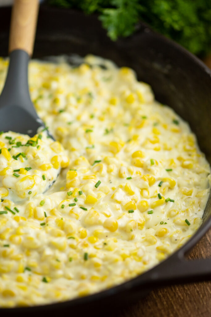 Homemade creamed corn in a large cast iron skillet with a wooden spoon in the middle topped with freshly chopped chives