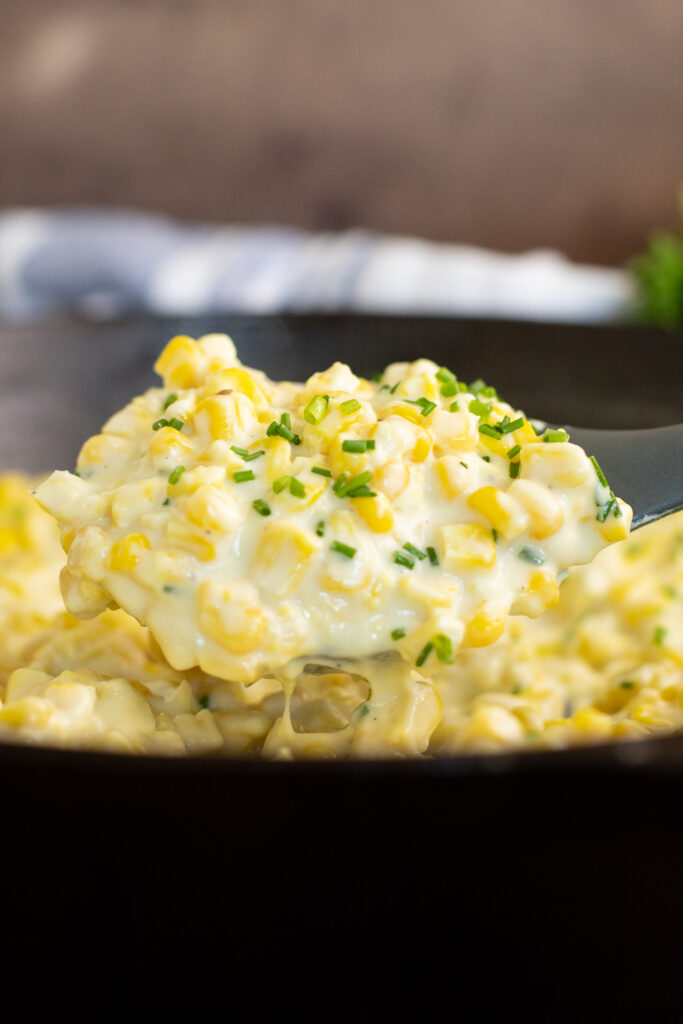 Homemade creamed corn topped with freshly chopped chives being scooped out of a cast iron skillet with a silicone spoon