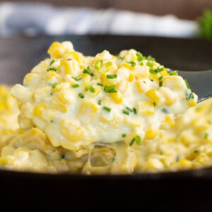 Homemade creamed corn topped with freshly chopped chives being scooped out of a cast iron skillet with a silicone spoon