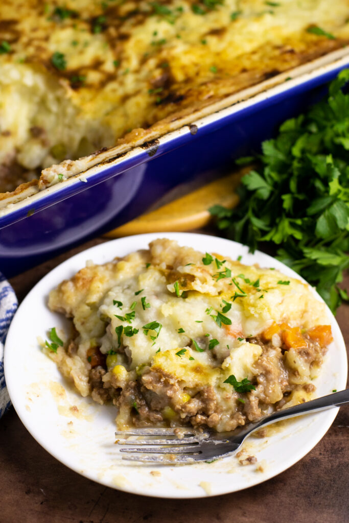 Serving of shepherd's pie on a small plate next to a fork with a bunch of parsley behind and the remaining casserole in a rectangular large dish