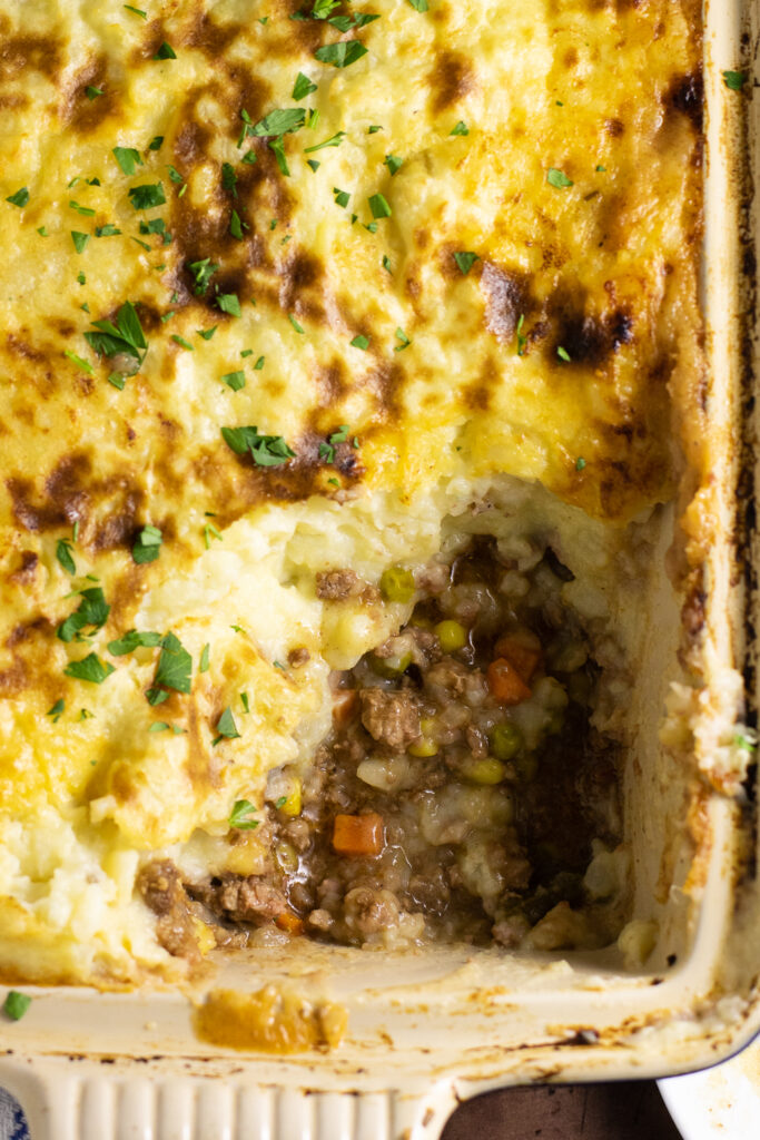 Close up overhead view of Shepherd's pie in a casserole dish topped with freshly chopped parsley with a scoop missing exposing the meat, vegetable, and gravy filling.