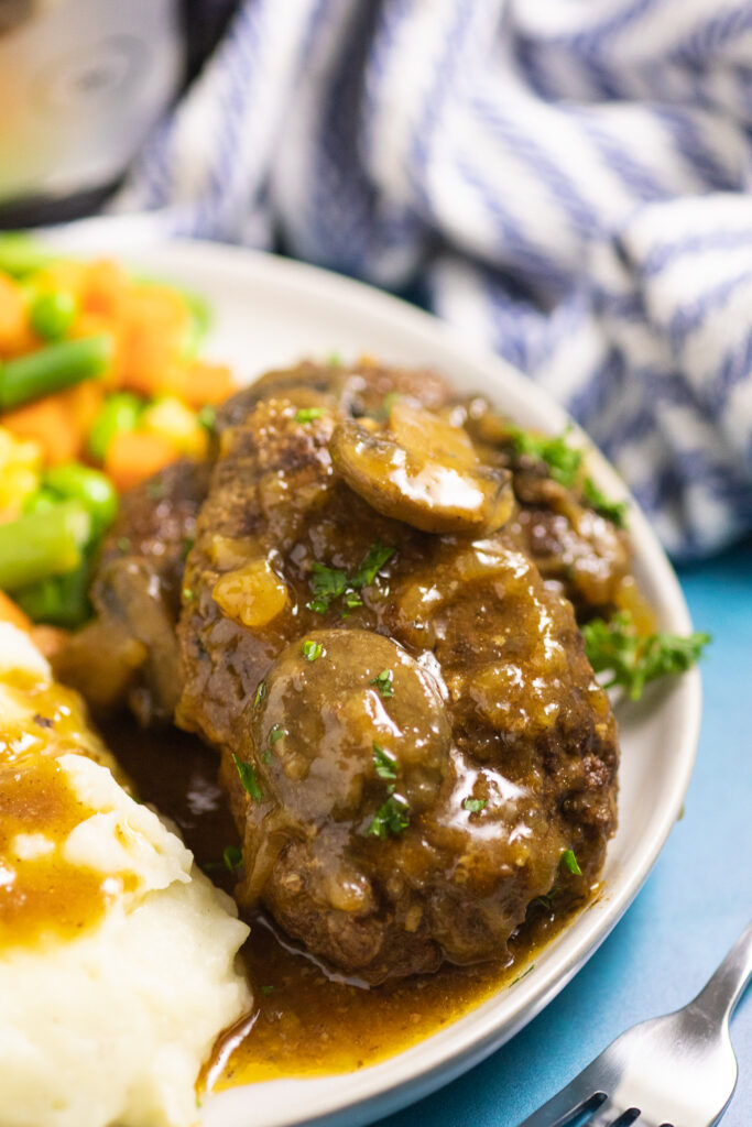 Slow cooker salisbury steak topped with mushroom and onion gravy and fresh chopped parsley on a small white plate with mashed potatoes and gravy and mixed vegetables with a striped linen in the background
