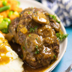 Slow cooker salisbury steak topped with mushroom and onion gravy and fresh chopped parsley on a small white plate with mashed potatoes and gravy and mixed vegetables