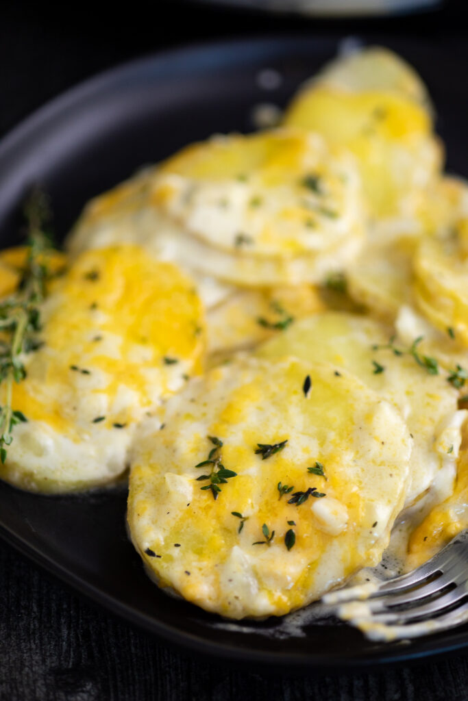 Close up of cheesy scalloped potatoes on a black plate next to a fork, topped with fresh thyme