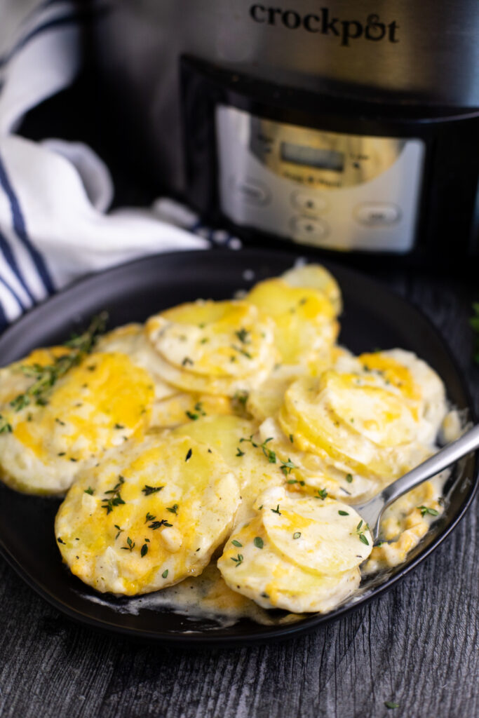 Slow Cooker scalloped potatoes topped with fresh parsley on a plate next to a fork in front of a Crock Pot on a rustic wood background