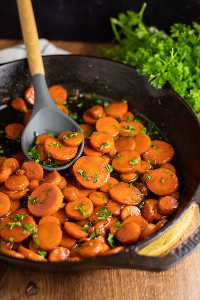Bourbon Glazed Carrots in a cast iron skillet topped with fresh chopped parsley with a wooden spoon in it. The skillet is on a rustic wood backdrop with a bunch of parsley behind.