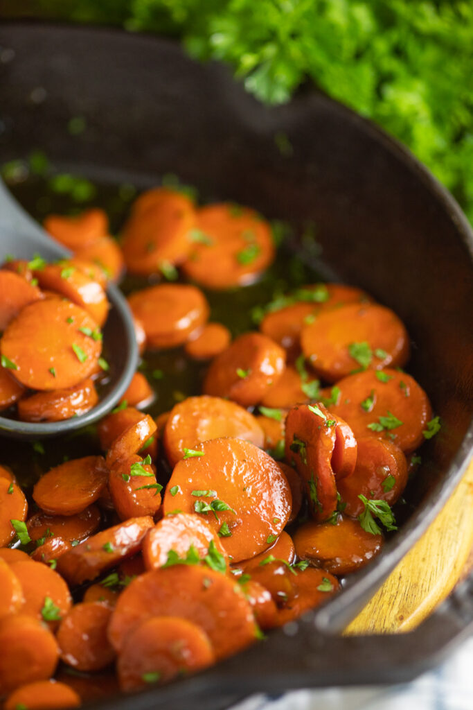 Close up of Bourbon Glazed Carrots cut into coins in a cast iron skillet on a rustic wood background with parsley behind