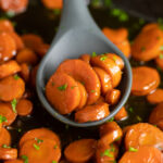 Carrots cut into coins in a brown sugar bourbon sauce in a silicone spoon resting in a cast iron skillet