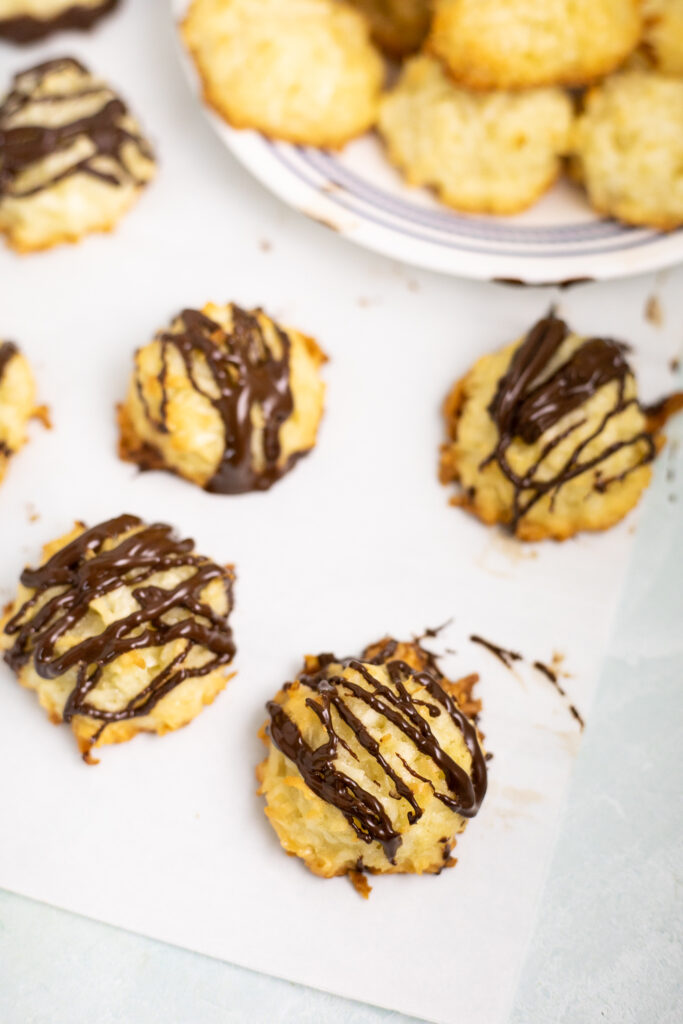 Four coconut macaroons with chocolate drizzled on top sitting on parchment paper with a small plate of plain coconut macaroons sitting behind