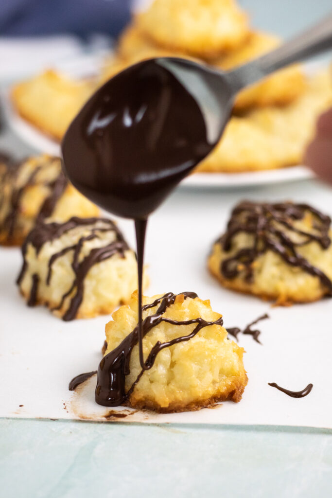 Spoon drizzling melted chocolate over a golden coconut macaroon with 2 more macaroons behind.