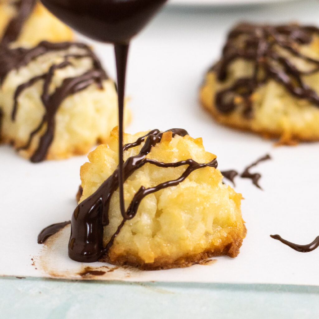 Spoon drizzling melted chocolate over a golden coconut macaroon with 2 more macaroons behind.