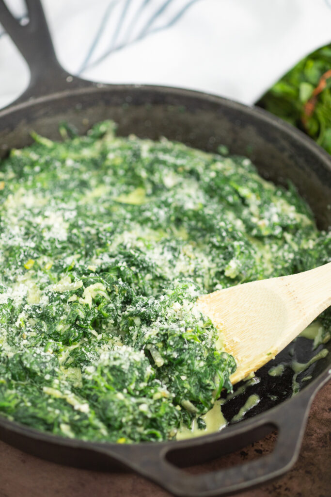 Close up of creamed spinach in a large cast iron skillet topped with freshly grated Parmesan cheese and a wooden spoon sticking out with a linen napkin and a bunch of fresh parsley behind it.
