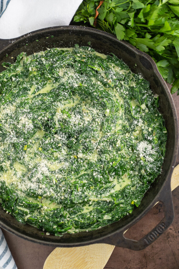 Overhead view of creamed spinach in a large cast iron skillet topped with freshly grated Parmesan cheese sitting next to a linen napkin, wooden spoon, and a bunch of fresh parsley.