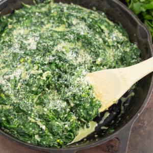 Close up of creamed spinach in a large cast iron skillet topped with freshly grated Parmesan cheese and a wooden spoon sticking out.