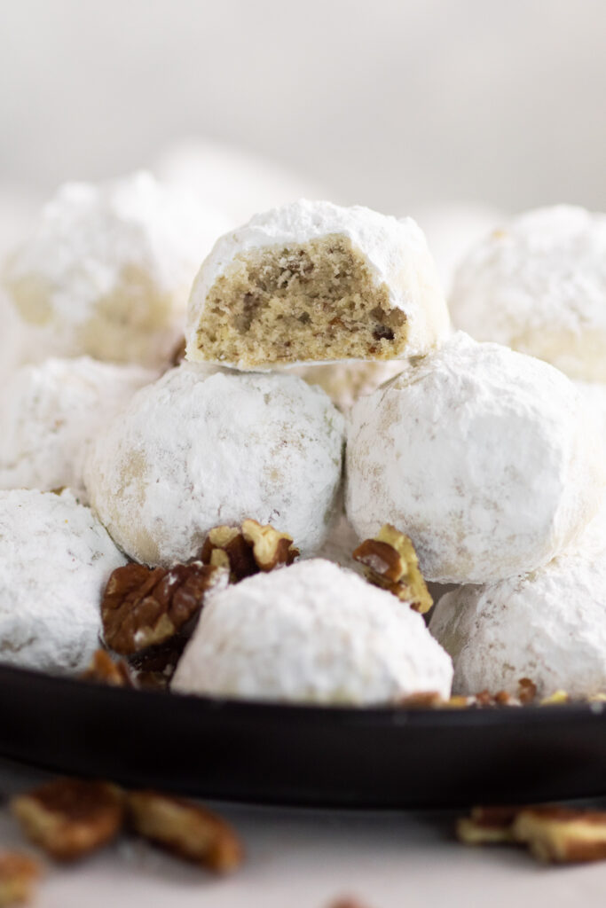 Pecan snowball cookies stacked on a plate. The top one has a bite taken out, and there are chopped pecans scattered with the cookies and on the white marble countertop.