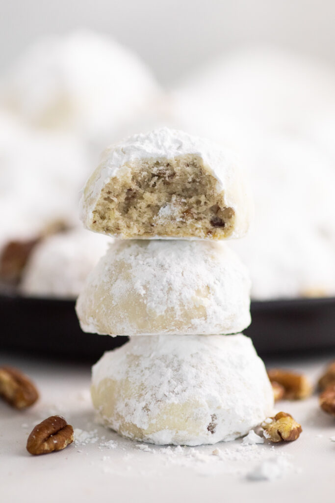 Stack of three pecan snowball cookies with a bite taken out of the top one, surrounded by chopped pecans with more cookies piled on a plate behind.