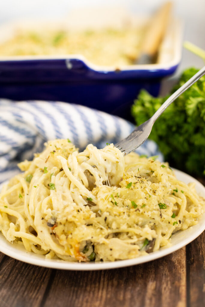A small plate piled high with turkey tetrazzini with a fork scooping up a bite. There is a linen napkin, a bunch of fresh parsley, and a large casserole in the background, all on a rustic wooden backdrop.