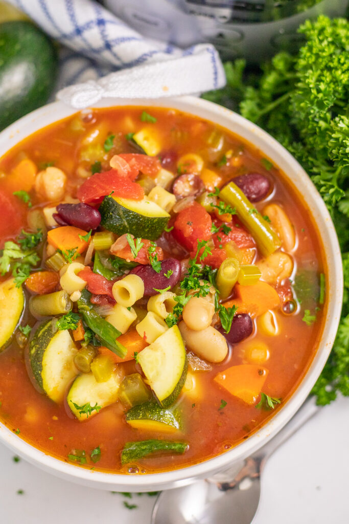Overhead view of slow cooker minestrone soup topped with fresh parsley on a white marble background next to a linen napkin and a spoon.