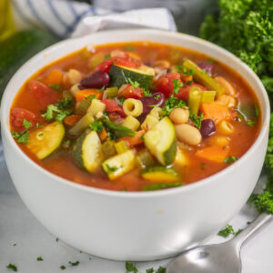 A white matte bowl full of slow cooker minestrone soup topped with fresh parsley, next to a spoon, a bunch of fresh parsley, and a blue and white striped linen napkin on a white marble background.