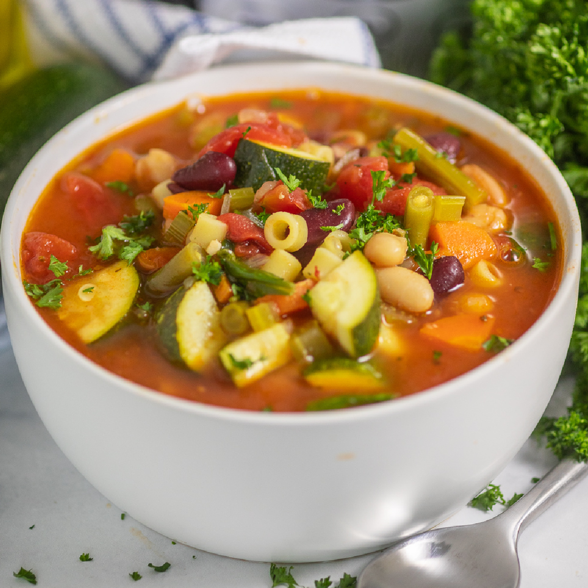 A white matte bowl full of slow cooker minestrone soup topped with fresh parsley, next to a spoon, a bunch of fresh parsley, and a blue and white striped linen napkin on a white marble background.