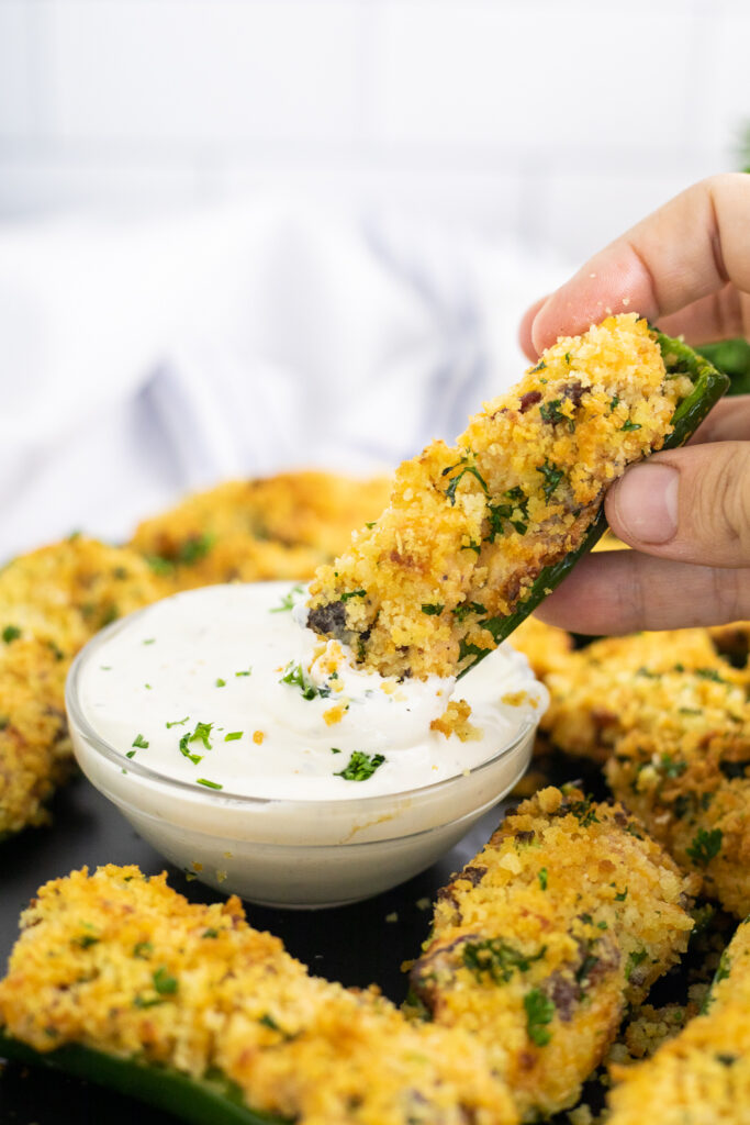 An air fryer jalapeno popper being dipped into a small glass bowl of ranch dressing surrounded by more poppers.